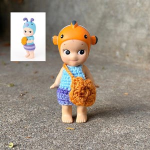 May include: A small figurine wearing an orange clownfish-themed headpiece, a light blue crocheted top, and purple crocheted shorts. The figure carries a small, round, orange crocheted bag across its body. A small inset image shows a similar figure.