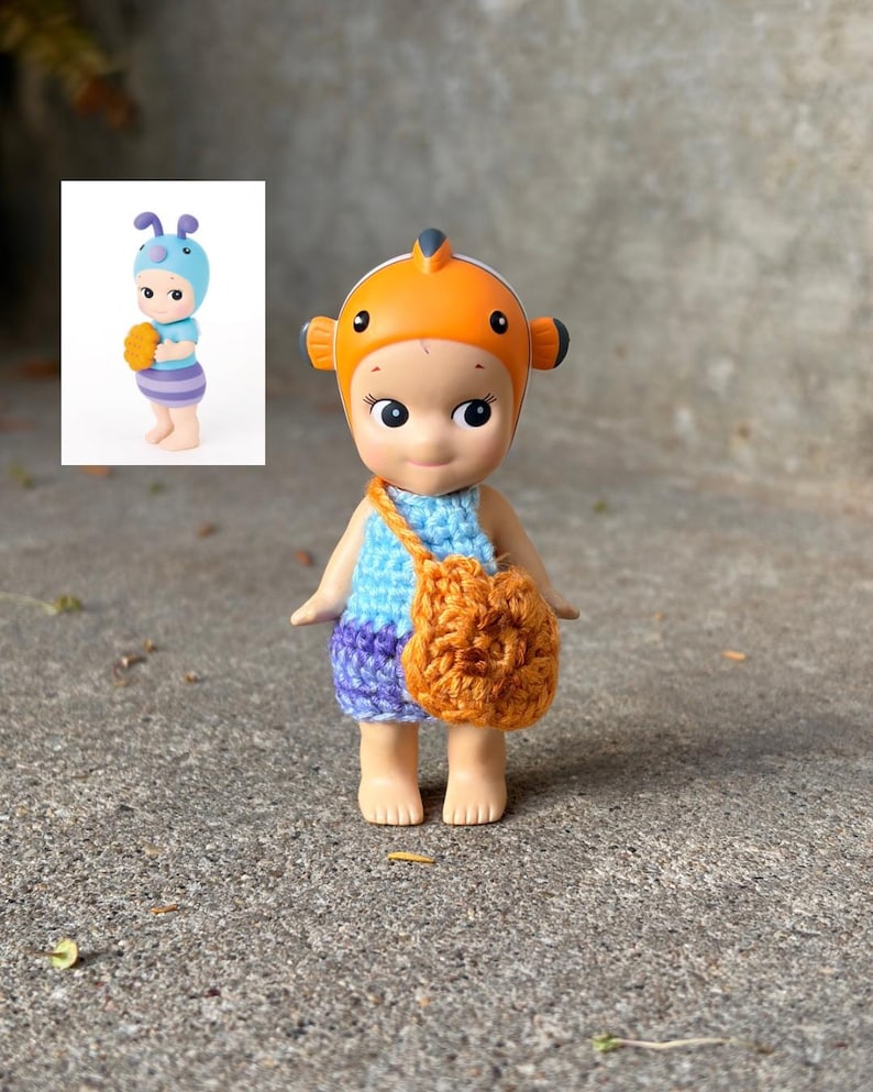 May include: A small figurine wearing an orange clownfish-themed headpiece, a light blue crocheted top, and purple crocheted shorts. The figure carries a small, round, orange crocheted bag across its body. A small inset image shows a similar figure.