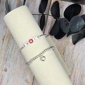 May include: Two bracelets are displayed. The top bracelet is silver with white letter beads spelling "OHIO" with a red heart. The bottom bracelet is silver with small beads and a heart charm. The bracelets are on a cream-colored cylindrical display.
