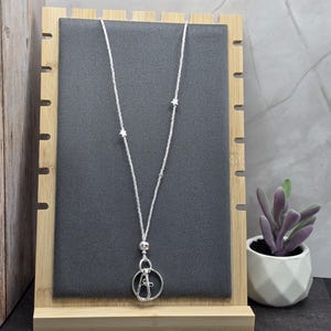 May include: A silver-colored necklace with a beaded chain and a pendant featuring a circular design with a letter "A" inside. The necklace is displayed on a black surface, set against a wooden frame.