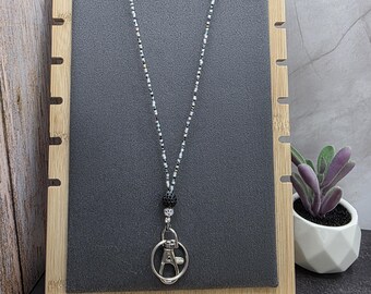 May include: A beaded lanyard with a silver-tone clasp and a black and silver bead design. The lanyard is displayed on a dark gray background, mounted on a bamboo display stand. A small potted plant is visible in the background.