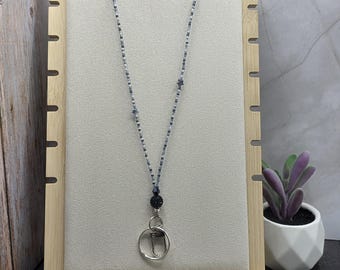 May include: A beaded lanyard with a silver-tone clasp and a dark blue beaded accent. The lanyard is displayed on a light beige background, set within a wooden frame. A small potted succulent is visible in the background.