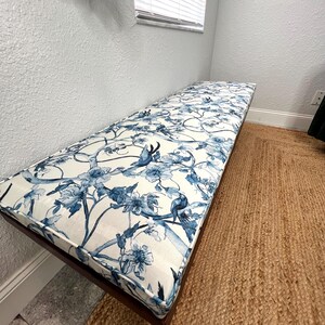 Peut inclure: Banc rectangulaire avec un cadre en bois fonc&eacute; et un coussin &agrave; motifs floraux. Le coussin pr&eacute;sente un fond blanc avec des motifs floraux et d'oiseaux bleus. Le banc est pos&eacute; sur un tapis tiss&eacute; marron.