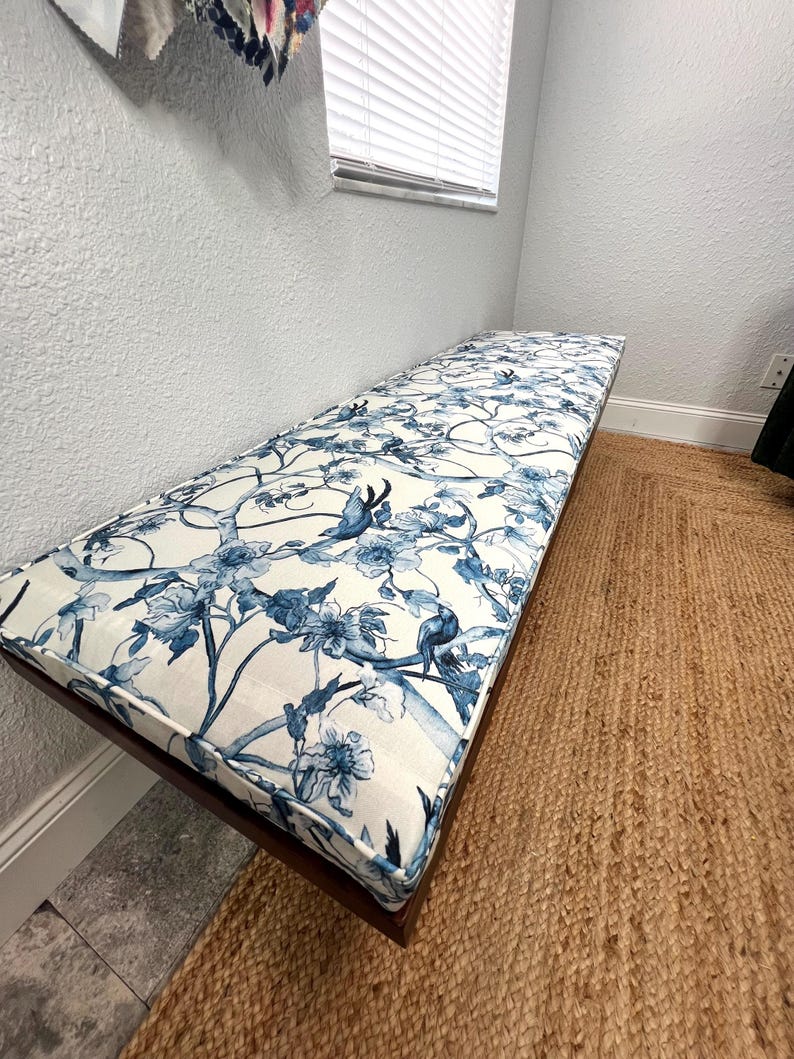 Peut inclure: Banc rectangulaire avec un cadre en bois fonc&eacute; et un coussin &agrave; motifs floraux. Le coussin pr&eacute;sente un fond blanc avec des motifs floraux et d'oiseaux bleus. Le banc est pos&eacute; sur un tapis tiss&eacute; marron.