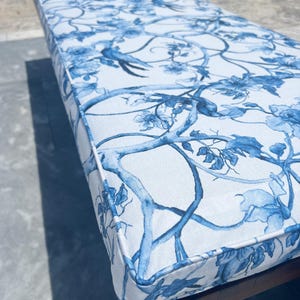 Peut inclure: Un coussin rectangulaire d'ext&eacute;rieur avec un fond blanc et un motif floral et d'oiseaux bleu. Le coussin a une bordure blanche et est plac&eacute; sur un cadre en bois. Le motif pr&eacute;sente des branches, des fleurs et des oiseaux.