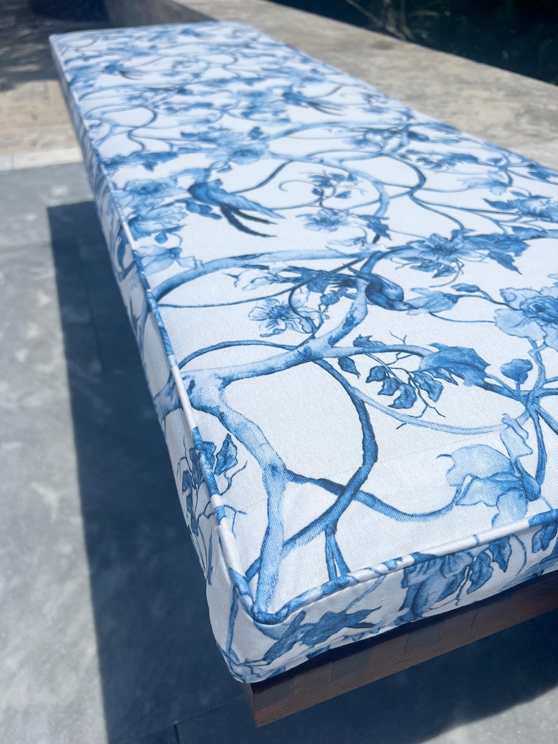 Peut inclure: Un coussin rectangulaire d'ext&eacute;rieur avec un fond blanc et un motif floral et d'oiseaux bleu. Le coussin a une bordure blanche et est plac&eacute; sur un cadre en bois. Le motif pr&eacute;sente des branches, des fleurs et des oiseaux.