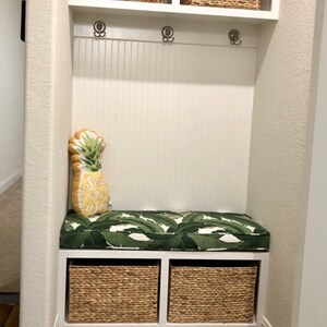 May include: A white entryway bench with a green and white leaf-patterned cushion and a pineapple-shaped pillow that says "Sea the Day." Two woven baskets sit below the bench. Above the bench are three coat hooks and two woven baskets.
