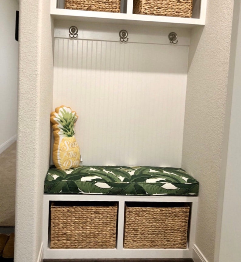 May include: A white entryway bench with a green and white leaf-patterned cushion and a pineapple-shaped pillow that says "Sea the Day." Two woven baskets sit below the bench. Above the bench are three coat hooks and two woven baskets.