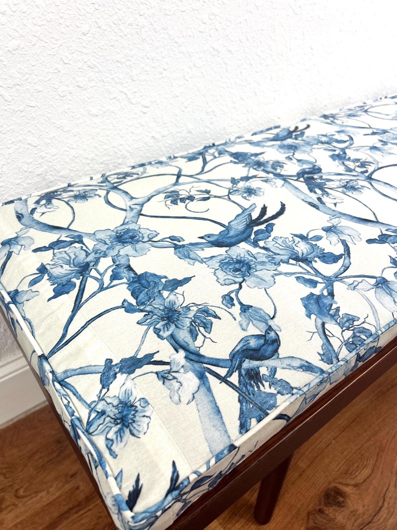 Peut inclure: Banc en bois avec un dessus rembourr&eacute; orn&eacute; d'un motif floral et d'oiseaux bleu et blanc. Le banc a un cadre et des pieds marron fonc&eacute;. Le coussin a un bord passepoil&eacute;.