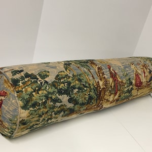 May include: A long, cylindrical bolster pillow with a floral print featuring a scene of people walking through a forest. The fabric is a mix of green, blue, and brown tones with red accents.