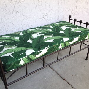 May include: A metal bench with a cushion featuring a green and white tropical leaf pattern. The bench has a dark brown metal frame with decorative finials on the backrest. The bench is approximately 48 inches long.