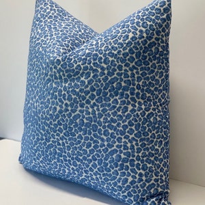 May include: A square decorative pillow with a blue and white spotted pattern. The pillow has a textured appearance and is placed on a white surface. The pillow is a home decor item.
