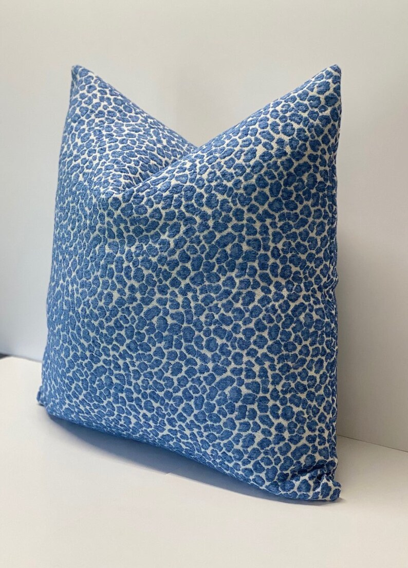 May include: A square decorative pillow with a blue and white spotted pattern. The pillow has a textured appearance and is placed on a white surface. The pillow is a home decor item.