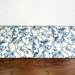 Peut inclure: Banc rectangulaire avec un motif floral et d'oiseaux bleu et blanc. Le motif pr&eacute;sente des fleurs et des oiseaux stylis&eacute;s sur un fond cr&egrave;me. Le banc est pos&eacute; contre un mur blanc et un sol en bois. Le banc mesure environ 120 cm de long.