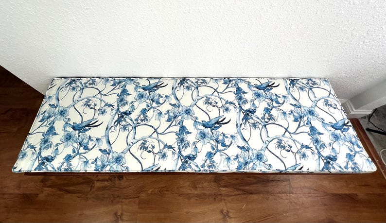 Peut inclure: Banc rectangulaire avec un motif floral et d'oiseaux bleu et blanc. Le motif pr&eacute;sente des fleurs et des oiseaux stylis&eacute;s sur un fond cr&egrave;me. Le banc est pos&eacute; contre un mur blanc et un sol en bois. Le banc mesure environ 120 cm de long.