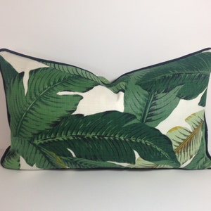 May include: Rectangular decorative pillow with a white background and a vibrant green banana leaf pattern. The pillow has a dark blue trim around the edges, adding a contrasting detail to the tropical design.