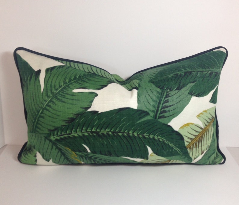 May include: Rectangular decorative pillow with a white background and a vibrant green banana leaf pattern. The pillow has a dark blue trim around the edges, adding a contrasting detail to the tropical design.