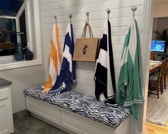 May include: A white bench with a blue and white zebra-striped cushion. Beach towels in orange, blue, black and white, and green hang on hooks above the bench. A brown tote bag with a seahorse graphic hangs on a hook.