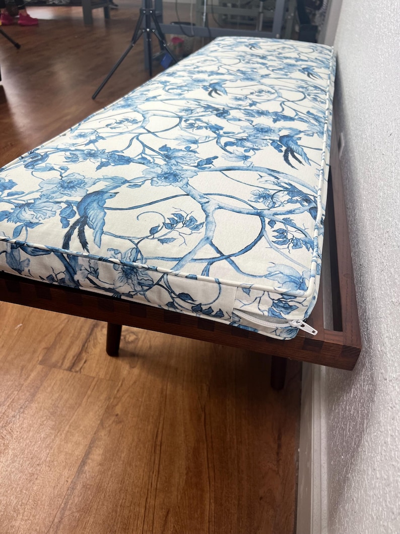 Peut inclure: Banc en bois long avec un coussin orn&eacute; d'un motif floral et d'oiseaux bleu et blanc. Le banc a un cadre et des pieds marron fonc&eacute;. Le coussin a une fermeture &eacute;clair sur le bord.