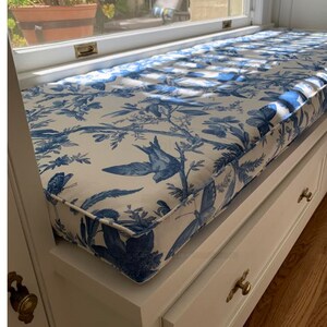 Peut inclure: Banquette blanche avec coussin en tissu toile de Jouy bleu et blanc, orn&eacute; d'oiseaux et de papillons. Le coussin repose sur un meuble de rangement blanc avec des poign&eacute;es en laiton. La banquette est pr&egrave;s d'une fen&ecirc;tre.