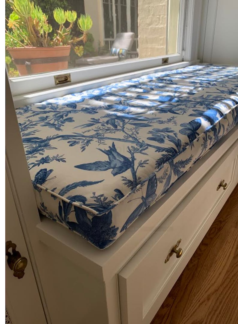 Peut inclure: Banquette blanche avec coussin en tissu toile de Jouy bleu et blanc, orn&eacute; d'oiseaux et de papillons. Le coussin repose sur un meuble de rangement blanc avec des poign&eacute;es en laiton. La banquette est pr&egrave;s d'une fen&ecirc;tre.