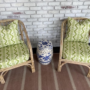 May include: Two rattan armchairs with green patterned cushions and pillows. A blue and white ceramic garden stool sits between the chairs. The chairs are on a striped rug, set against a white brick wall.