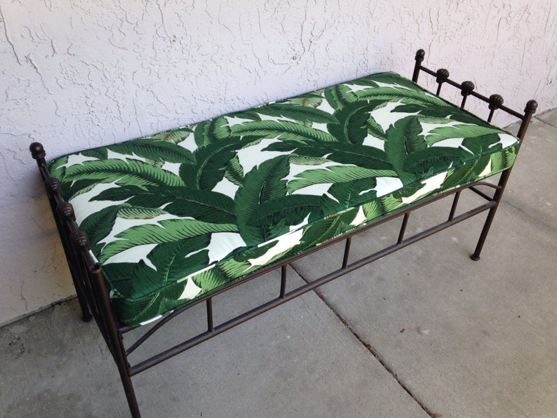 May include: A metal bench with a rectangular cushion. The cushion features a white background with a pattern of large green leaves. The metal frame is dark brown with decorative finials at the ends of the backrest.