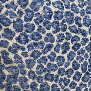 May include: Close-up of a fabric with a blue leopard print pattern on a cream background. The pattern features irregular, rounded shapes in varying shades of blue, creating a textured appearance. The fabric appears to be soft and possibly used for upholstery or home decor.