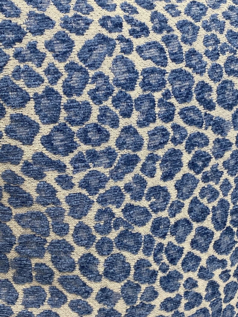 May include: Close-up of a fabric with a blue leopard print pattern on a cream background. The pattern features irregular, rounded shapes in varying shades of blue, creating a textured appearance. The fabric appears to be soft and possibly used for upholstery or home decor.