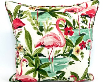 Peut inclure: Un coussin blanc avec un imprimé tropical représentant des flamants roses, des palmiers verts et des fleurs rouges et jaunes.