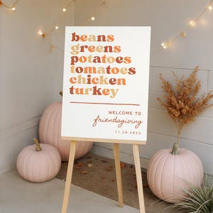 Puede incluir: Un letrero blanco de Friendsgiving sobre un caballete de madera, que enumera los elementos del menú en tonos naranja y marrón. Calabazas rosas y luces decorativas rodean el letrero. El letrero dice "Welcome to Friendsgiving 11.20.2025".
