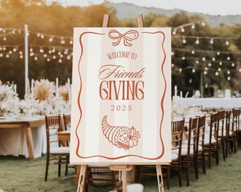 May include: A wooden easel displays a sign with the words "Welcome to Friends Giving 2025." The sign has a cream background with vertical stripes and a brown border. A bow and cornucopia graphic are at the top and bottom.