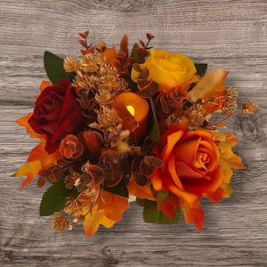 May include: A fall-themed floral arrangement featuring a central lit candle surrounded by red, orange, and yellow roses, along with autumn leaves and decorative sprigs. The arrangement is set against a wooden background.
