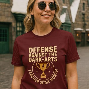 May include: A maroon t-shirt with the text "DEFENSE AGAINST THE DARK-ARTS" in gold lettering. Below the text is a gold trophy graphic with a lightning bolt. The shirt also says "TEACHER OF THE MONTH".