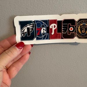 Peut inclure: Un autocollant avec le mot "DEPLC" en lettres majuscules, chaque lettre représentant une équipe sportive de Philadelphie. Les lettres sont aux couleurs des équipes : vert, bleu, rouge et noir. L'autocollant est tenu par une main avec du vernis à ongles rouge.