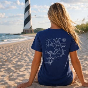 May include: Navy blue t-shirt featuring a white map design with coastal details and place names. The shirt is worn on a beach with a lighthouse in the background. The map includes a compass rose.