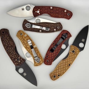 May include: An assortment of folding knives with various handle designs and blade finishes. The handles feature intricate patterns in shades of brown, red, and tan. The blades are silver and black, showcasing the Spyderco logo.