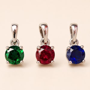 May include: Three gemstone pendants with silver-colored settings. One pendant features a round, emerald-green stone, another a ruby-red stone, and the third a sapphire-blue stone. The pendants are displayed against a light background.