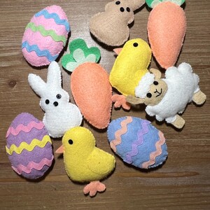Peut inclure: Ensemble de décorations de Pâques en feutrine, comprenant des œufs colorés, des lapins, des poussins, un agneau et des carottes. Les œufs sont décorés de motifs ondulés de différentes couleurs. Le lapin et l'agneau sont blancs, le poussin est jaune et la carotte est orange.