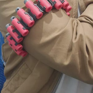 May include: A series of red and gray cylindrical objects, possibly ammunition or a prop, are arranged in a row. The objects are attached to a gray base. The items are resting on a tan jacket.