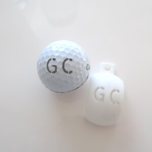 May include: A white golf ball and a white golf ball marker, both with the initials "GC" stenciled in gray. The golf ball has a dimpled surface. The marker is a small, rounded shape with a hole at the top.