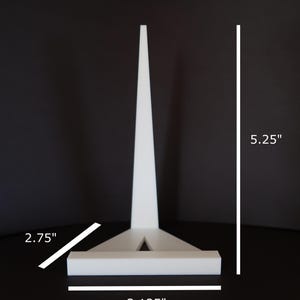 May include: A white, geometric, decorative object with a triangular base and a tall, pointed top. The object is 5.25 inches tall, 3.125 inches wide, and 2.75 inches deep. The background is a solid black.