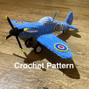 May include: A blue crochet airplane with black propeller and wheels. The wings and body feature roundels with red, white, and blue circles. The text "Crochet Pattern" is visible on the wooden surface.