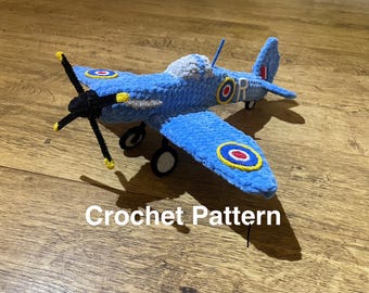 Puede incluir: Un avión azul de ganchillo con hélice y ruedas negras. Las alas y el cuerpo presentan escarapelas con círculos rojos, blancos y azules. El texto "Crochet Pattern" es visible.
