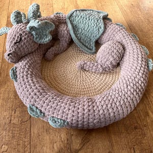 Puede incluir: Una cama para mascotas de drag&oacute;n hecha a mano con ganchillo en tonos lavanda y azul claro. El drag&oacute;n est&aacute; enroscado en forma circular, con un centro beige. El drag&oacute;n tiene los ojos cerrados. La cama est&aacute; sobre una superficie de madera.