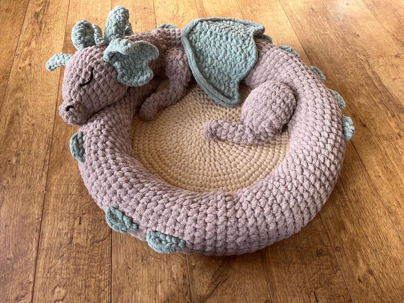 Puede incluir: Una cama para mascotas de drag&oacute;n hecha a mano con ganchillo en tonos lavanda y azul claro. El drag&oacute;n est&aacute; enroscado en forma circular, con un centro beige. El drag&oacute;n tiene los ojos cerrados. La cama est&aacute; sobre una superficie de madera.
