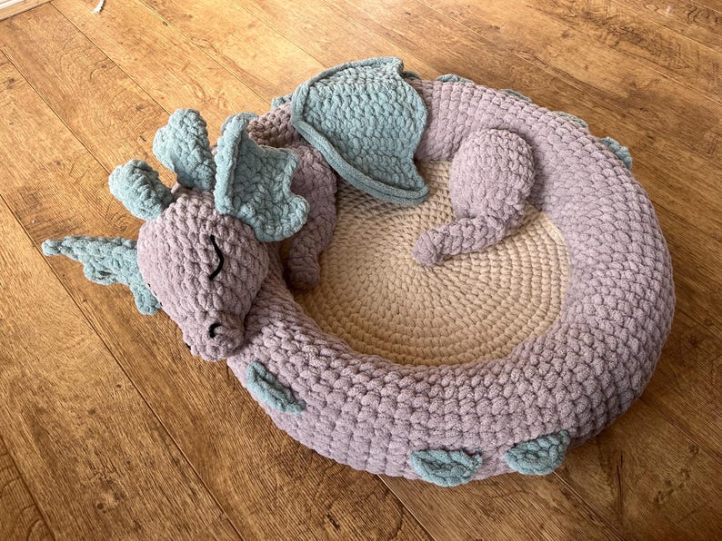 Puede incluir: Una cama para mascotas de drag&oacute;n hecha a mano, tejida a ganchillo, en forma circular. El drag&oacute;n es de color lavanda con detalles en azul claro en las alas y las orejas. El centro de la cama es de color beige claro, creando un espacio acogedor.
