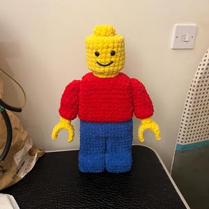 May include: A handmade, crocheted Lego figure in primary colors. The figure has a yellow head, red torso, blue legs, and yellow hands. The face has a simple, smiling expression. The figure is approximately 12 inches tall.