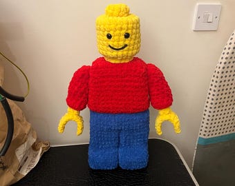 Puede incluir: Una figura de Lego hecha a mano, tejida a ganchillo, en colores primarios. La figura tiene una cabeza amarilla, un torso rojo, piernas azules y manos amarillas. La cara tiene una expresión simple y sonriente. La figura mide unos 30 cm de alto.
