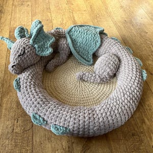 Puede incluir: Una cama para mascotas con forma de drag&oacute;n, tejida a ganchillo en tonos grises y turquesa. El drag&oacute;n est&aacute; enroscado, con la cabeza apoyada en su cuerpo. La cama tiene forma circular, con un di&aacute;metro de unos 61 cm.